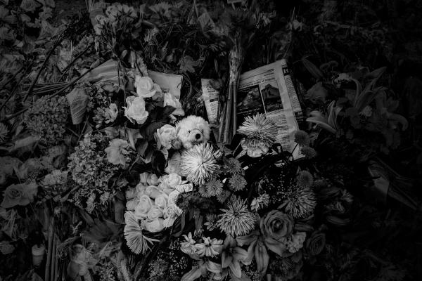 A dense cluster of memorial gifts, including flowers, newspapers, and a stuffed bear at the centre of the image.